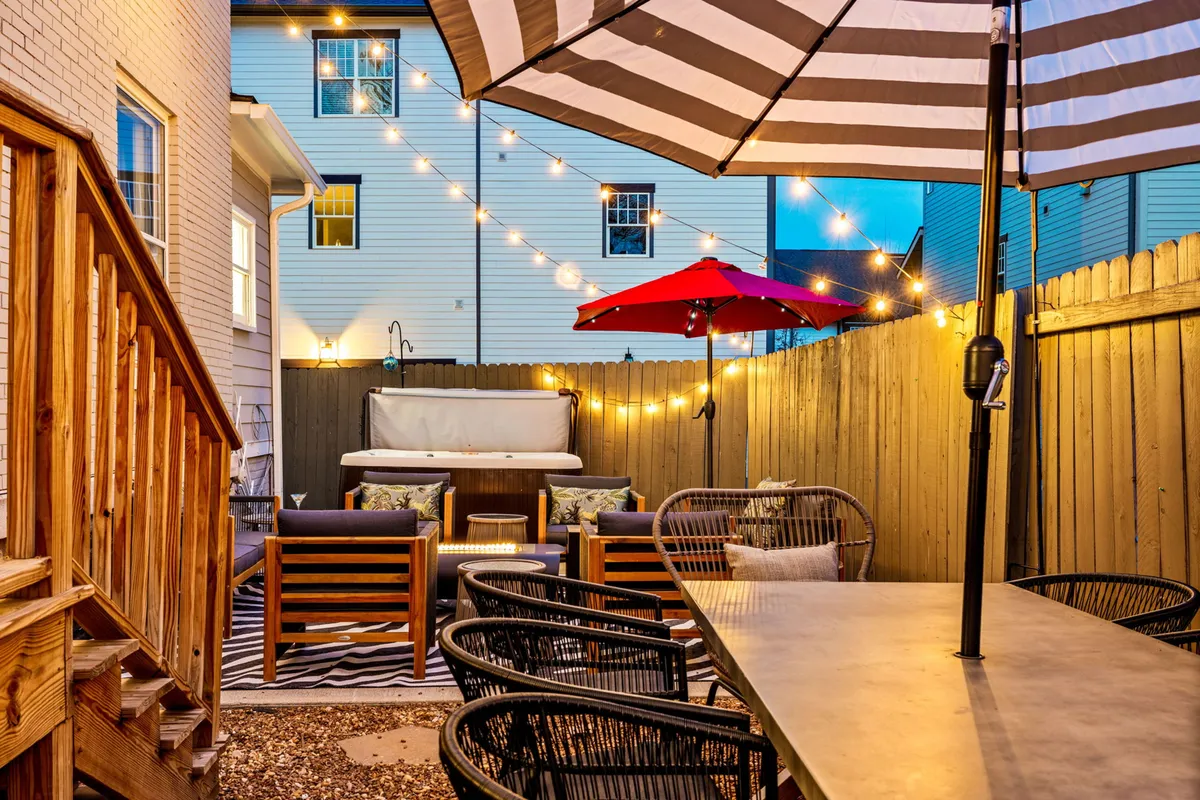 An inviting backyard scene featuring striped umbrellas, festive string lights, a private hot tub, and a comfortable seating area arranged for conversation or relaxation. The area radiates both privacy and the ambiance of a lively gathering spot.