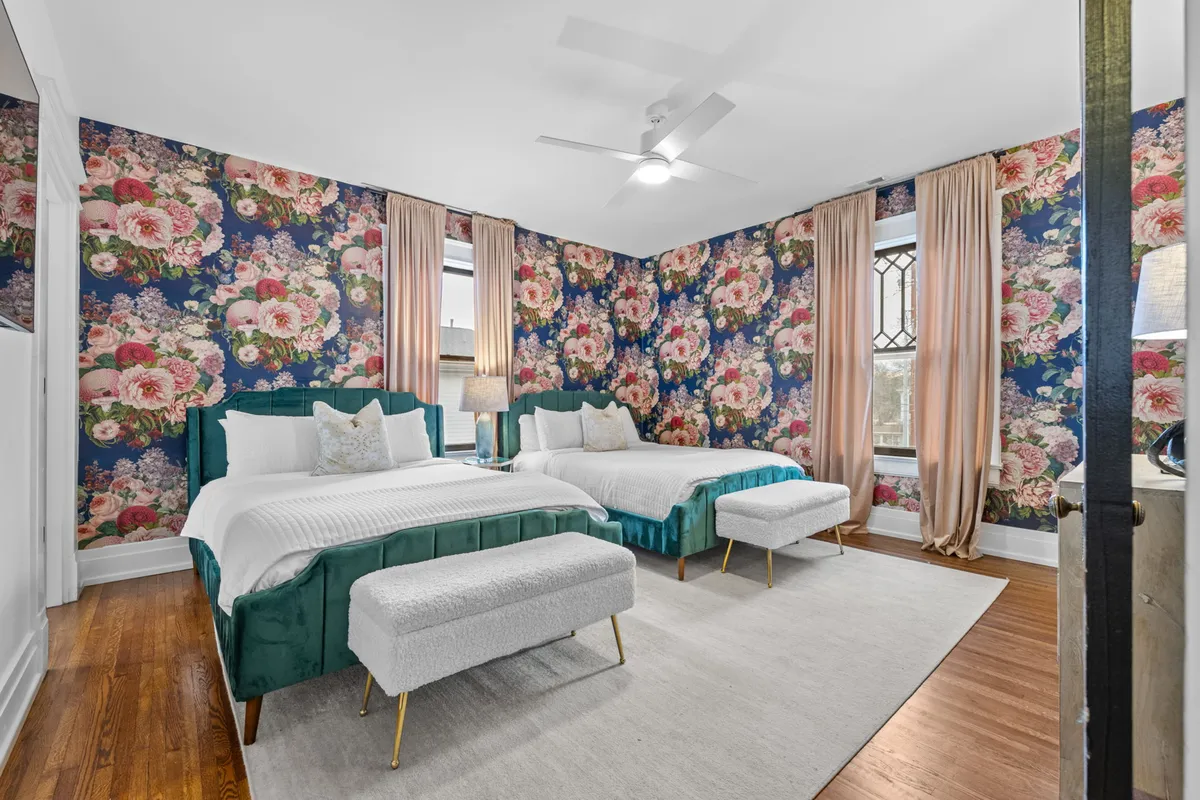 A sunlit bedroom with large windows and cheerful floral wallpaper. The bed features teal accents and is complemented by a cozy bench at the foot, while curtains frame the windows, inviting natural light into the space.