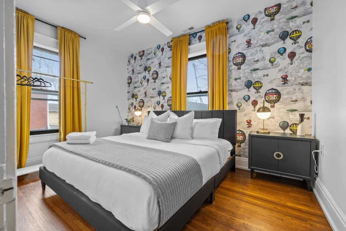 A whimsical bedroom with colorful hot air balloon wallpaper, a queen-sized bed, yellow curtains, and nightstands with lamps. The decor exudes comfort and playful charm, making the room feel both inviting and unique.