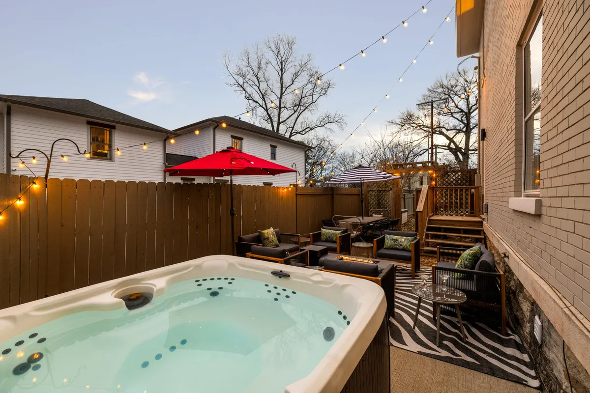 A cozy outdoor space with a bubbling hot tub, ample seating, and string lights draped overhead. The setup is ideal for group gatherings or intimate moments under the stars, with greenery providing a sense of seclusion.