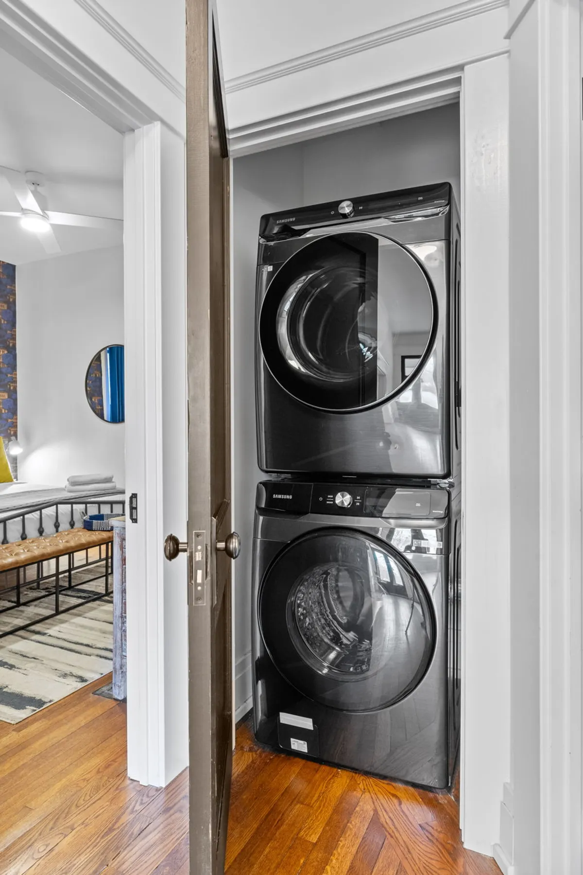 A stacked washer and dryer are positioned near an open door, with wooden flooring beneath. The scene is simple and functional, emphasizing cleanliness and modern convenience.