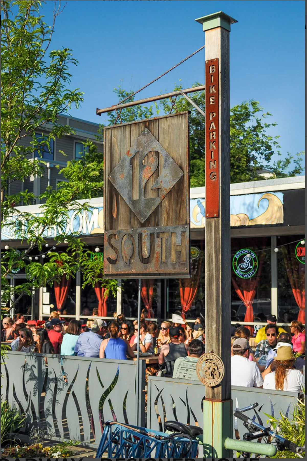 A vibrant exterior scene featuring the iconic '12 South' sign, bike parking, and a bustling outdoor dining area filled with people enjoying local cuisine. The atmosphere is lively and social, with friends gathering and people-watching under the shade of trees.