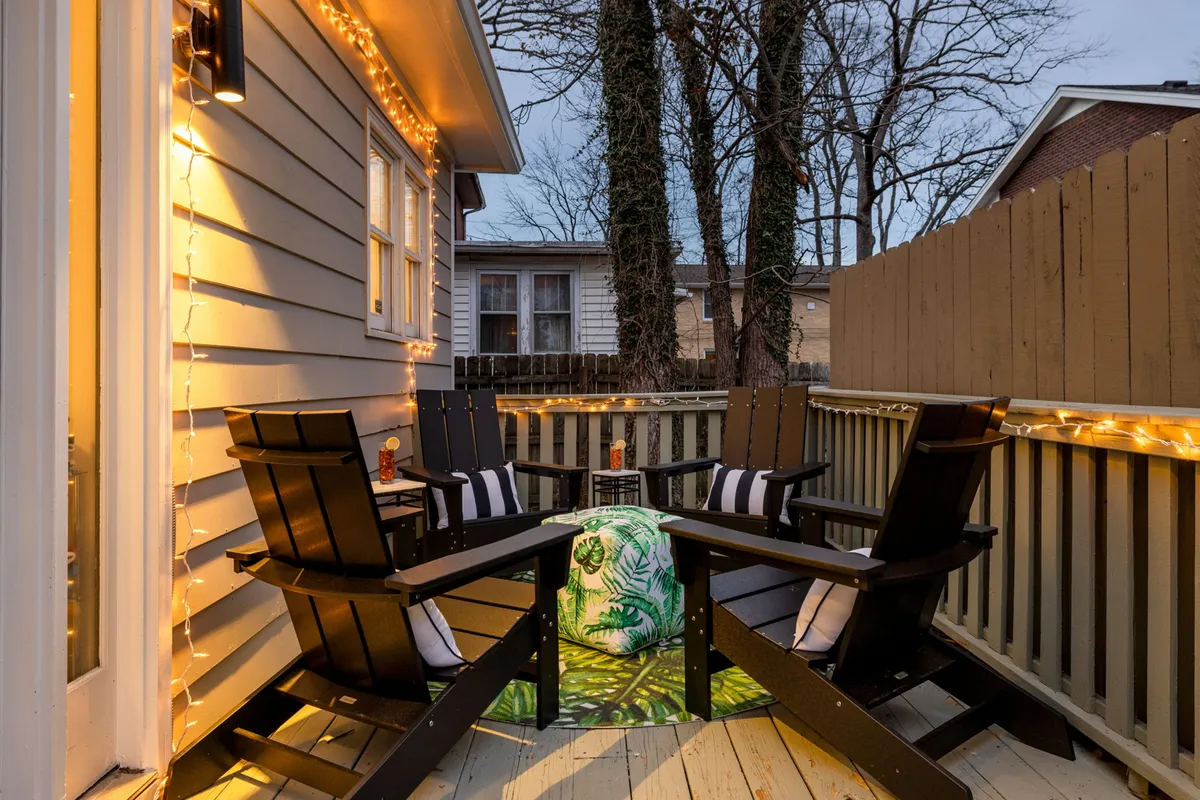A fire pit area framed by Adirondack chairs and illuminated by string lights in a fenced yard, evoking warmth and inviting conversation. This setup is designed for guests to enjoy evenings outdoors in comfort and privacy.