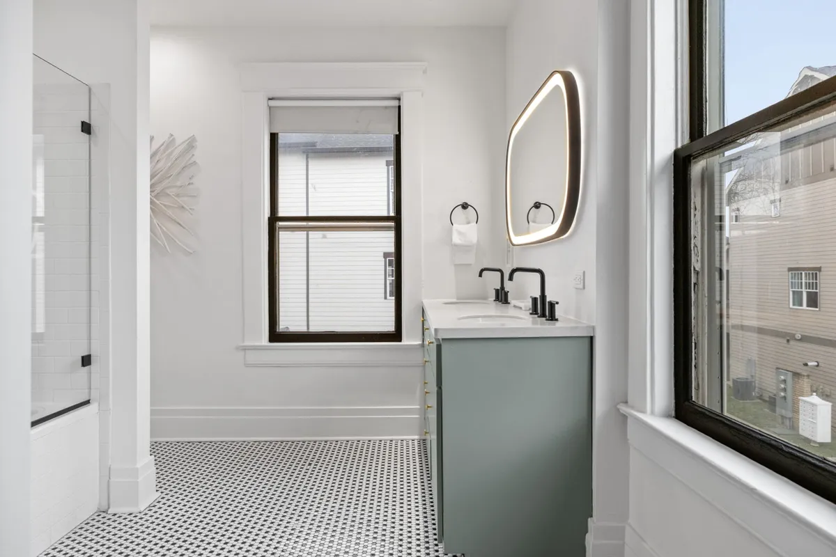 A modern bathroom featuring a dual-sink vanity, a large backlit mirror, and geometric patterned floor tiles. The space is serene and spacious, ideal for morning routines or relaxation after a long day.